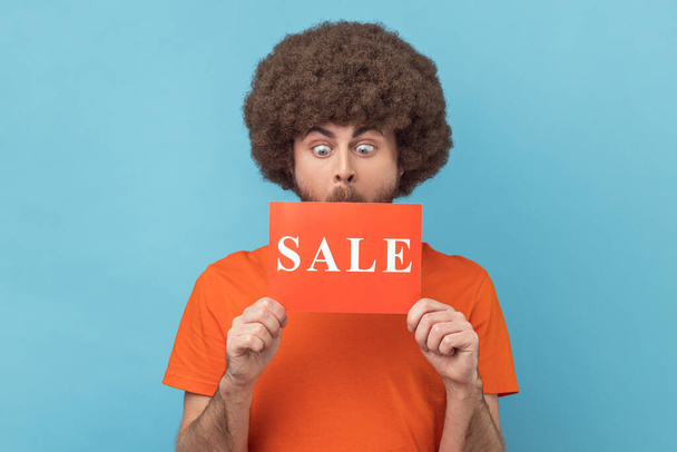 Man with Afro hairstyle wearing orange T-shirt standing with Sale word, looking with big eyes surprised, shocked by discounts, Black Friday low prices. Indoor studio shot isolated on blue background. - Zdjęcie, obraz