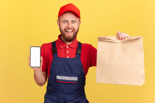 Joyful satisfied deliveryman holding paper parcel and showing smart phone with blank display for advertisement, looking at camera with smile. Indoor studio shot isolated on yellow background. - Valokuva, kuva