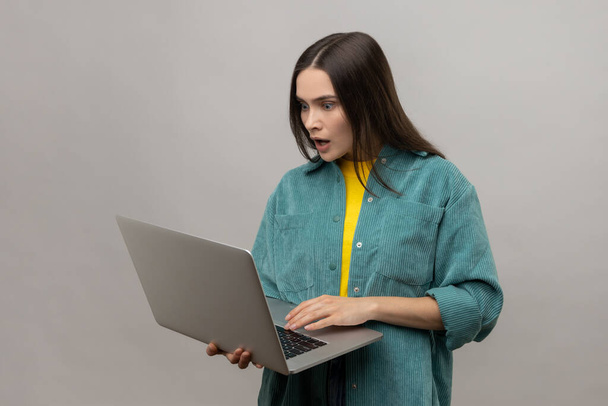 Portrait of shocked surprised young adult woman holding laptop and looking at display with big astonished eyes, wearing casual style jacket. Indoor studio shot isolated on gray background. - Photo, Image