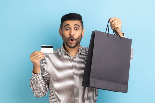 Astonished businessman holding and showing limitless credit card and shopping bags, surprised with great shopping, wearing striped shirt. Indoor studio shot isolated on blue background. - Zdjęcie, obraz