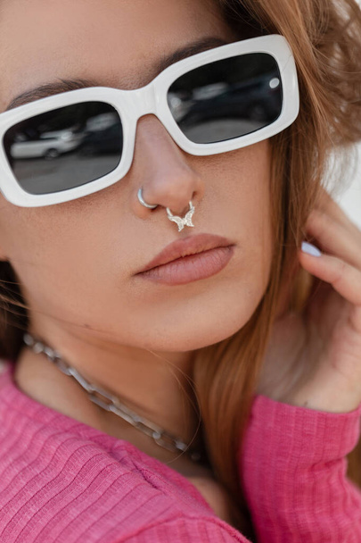 Fresh summer fashion close-up portrait of a stylish beautiful young woman with trendy white sunglasses with a nose piercing wearing a pink knit top. Clear female face - Fotó, kép