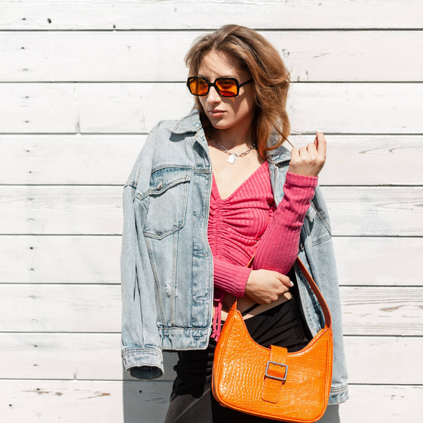 Moda bella donna elegante modello con occhiali da sole arancione brillante alla moda in eleganti vestiti jeans con una giacca e top rosa con una borsa di pelle arancione si trova vicino alla parete di legno bianco - Foto, immagini