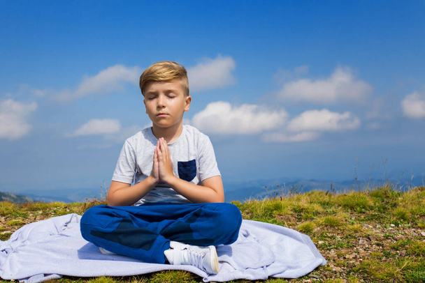 Спокойный мальчик с сцепленными руками практикует йогу с закрытыми глазами на вершине горы. Спокойный ребенок в намасте позирует против неба. - Фото, изображение