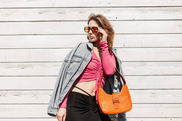 Cool fashion vogue beautiful model girl hipster wears orange sunglasses in fashion jeans jacket, pink top and black skirt walking near the vintage white wooden wall on the beach - Photo, Image