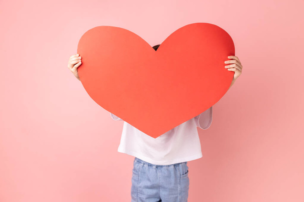 Portrait of anonymous little girl wearing white T-shirt hiding his face behind big red paper heart, making anonymous surprise, sending greeting card. Indoor studio shot isolated on pink background. - Foto, imagen