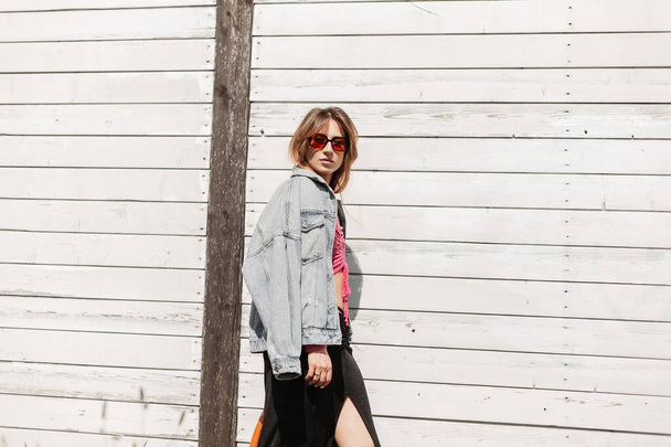 Menina bonita na moda em roupas de calça de primavera elegante com jaqueta de ganga e óculos de sol na moda andando perto de uma parede de madeira branca - Foto, Imagem
