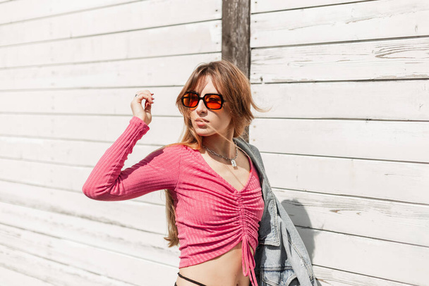 Sommer weiblichen Lebensstil Porträt einer stilvollen schönen jungen Frau mit modischer Sonnenbrille in einem trendigen rosa Crop Top und Jeansjacke zu Fuß am Strand in der Nähe einer hölzernen weißen Wand - Foto, Bild