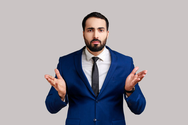 Portrait of bearded man standing with raised hands and confused indignant expression, asking why how, what reason, wearing official style suit. Indoor studio shot isolated on gray background. - Photo, image