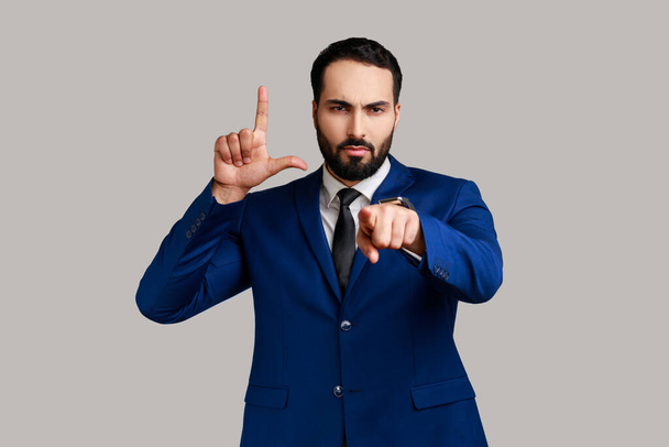 Portrait of strict bearded man pointing at camera and showing lame or loser gesture, mocking your failures, wearing official style suit. Indoor studio shot isolated on gray background. - Foto, immagini