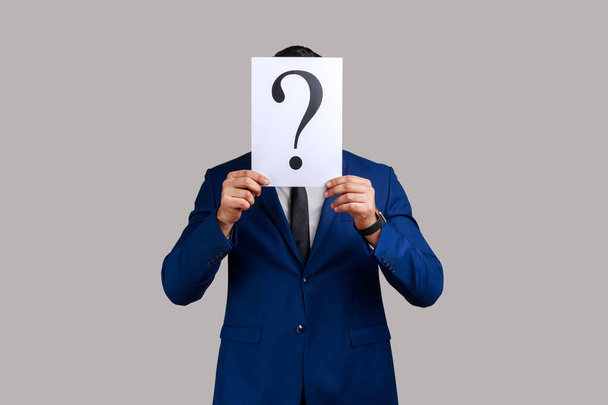 Unknown man hiding his face behind white paper with question mark, finding smart solution, asking for advice, wearing official style suit. Indoor studio shot isolated on gray background. - Foto, Bild