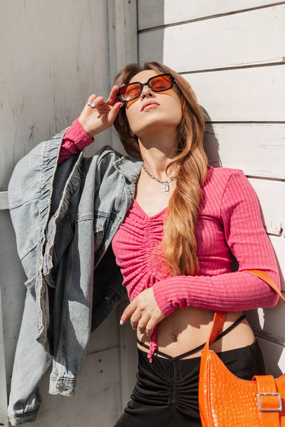 Modisch schöne Frau Modell mit cooler orangefarbener Sonnenbrille trägt ein rosafarbenes Stricktop mit Jeansjacke und Handtasche, die in der Nähe eines alten weißen Holzgebäudes steht - Foto, Bild