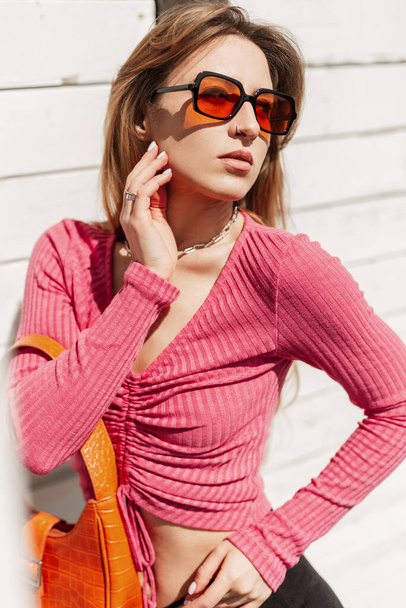 Stylish beautiful young redheaded girl with fashion orange sunglasses in fashionable bright summer clothes with a leather bag poses near a white wooden building on the beach - 写真・画像