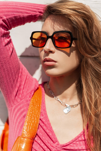 Elegante bella giovane donna con occhiali da sole arancio fresco, indossando abiti estivi alla moda e una borsa si erge e posa vicino a un muro di legno bianco in una giornata di sole - Foto, immagini