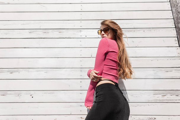 Modelo de menina elegante bonita e esbelta com óculos de sol na moda roupas de verão brilhantes com um top rosa e uma saia preta posa perto de uma parede de madeira branca - Foto, Imagem