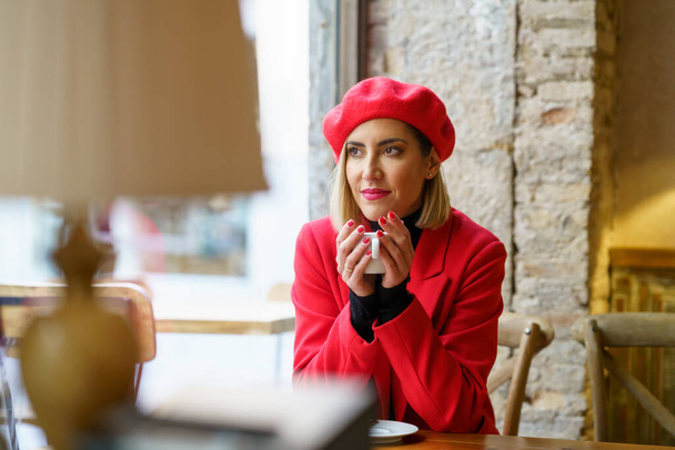 Piękna kobieta w czerwonym stroju odwracając wzrok marzycielską twarzą siedząc przy stole z filiżanką kawy w dłoniach w stołówce - Zdjęcie, obraz
