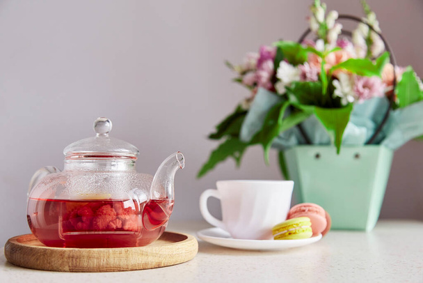 Útulný čaj čas - šálek čaje s makarony, skleněná konvice, svíčka a kytice květin. Bylinkový malinový přírodní čaj. Estetický domov s čajem a dezertem. Vlastní péče, relaxace, wellness životní styl - Fotografie, Obrázek