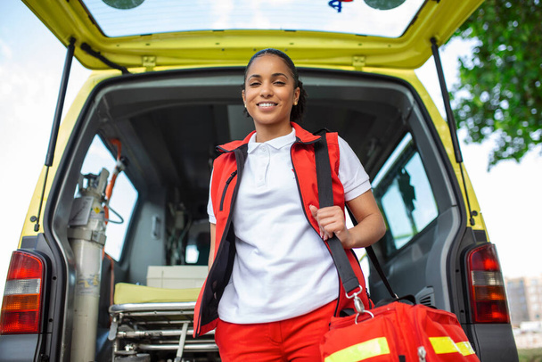 Die junge Frau, eine Rettungssanitäterin, steht im hinteren Teil eines Krankenwagens vor den offenen Türen. Sie blickt mit selbstbewusstem Gesichtsausdruck in die Kamera, lächelt und trägt eine medizinische Traumata-Tasche auf der Schulter.. - Foto, Bild