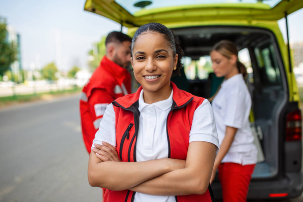 Молода жінка-афроамериканка посміхається, стоячи позаду машини швидкої допомоги, схрестивши руки. Три фельдшери на машині швидкої. - Фото, зображення