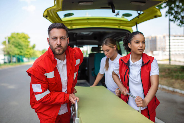 Junge Sanitäter ziehen in Eile Krankentransportwagen aus dem Auto. Sanitäter in Uniform tragen Trage aus dem Rettungswagen - Foto, Bild