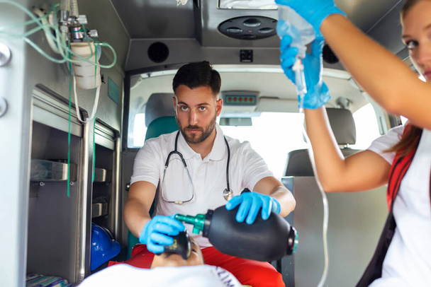 Equipe de paramédicos da EMS fornece ajuda médica ao paciente ferido a caminho do hospital de saúde. Assistente de Emergência Usando Máscara de Ventilação em Ambulância. Jovem enfermeira segurando solução IV - Foto, Imagem