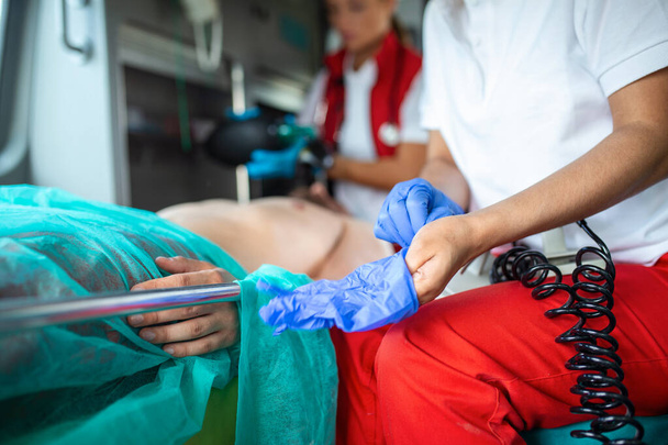 Afro-Amerikaanse paramedicus zet op Blue Surgical Rubber Handschoenen in een Ambulance Vehicle met een gewonde patiënt. Spoedeisende medische technicus controleert de vitale functies van het slachtoffer.. - Foto, afbeelding