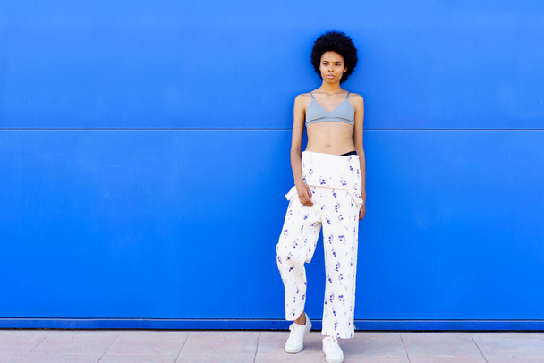 Full body of fit African American female on overall and bra looking away while standing on blue background in city - Foto, Imagem