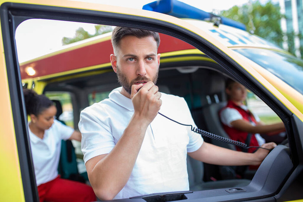 komea mies ensihoitaja puhuu kannettavalla radiolla ja seisoo lähellä ambulanssia
 - Valokuva, kuva