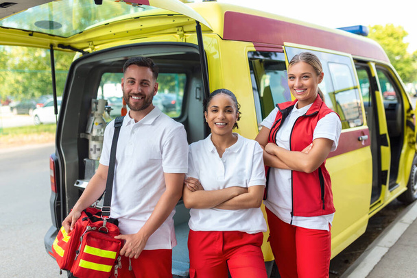 Kolme monirotuista ensihoitajaa seisoo ambulanssin ajoneuvon edessä kantaen kannettavia laitteita - Valokuva, kuva