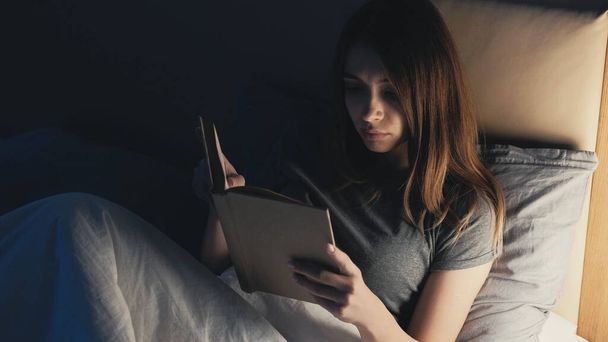 Night reading. Home leisure. Rest time. Relaxed woman lying in bed with interesting book turning off light for sleep in dark room interior. - Foto, Imagen