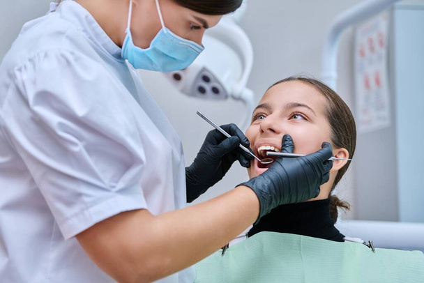 Young teenage female at dental checkup in clinic. Teenage girl sitting in chair, doctor dentist with tools examining patients teeth. Adolescence, hygiene, treatment, dental health care - Foto, imagen