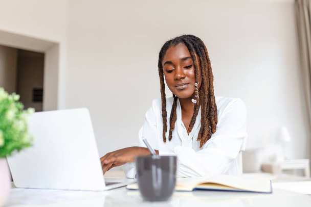 Belle femme africaine pigiste notant des informations pour la planification de projet faisant travail à distance via ordinateur portable. femme riant tout en lisant l'e-mail sur l'ordinateur portable moderne . - Photo, image