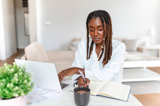 Concepto de trabajo remoto, tecnología y personas: joven mujer de negocios africana con computadora portátil y papeles trabajando en la oficina en el hogar durante la crisis de salud de Covid-19. - Foto, Imagen
