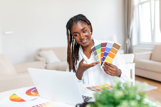 Vidám afro-amerikai női tervező videokonferenciát tart ügyfeleivel, asztalnál ül a számítógép előtt, színes palettákat tart, gesztikulál és mosolyog, másolja a helyet - Fotó, kép
