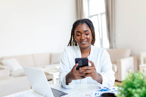 若い笑顔アフリカのビジネス女性がオフィスでコンピュータの近くでスマートフォンを使用して - 写真・画像