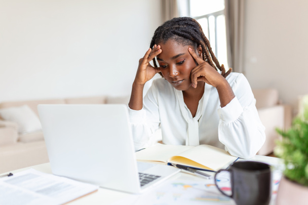 Una mujer de negocios agotada con dolor de cabeza en su oficina. Mujer creativa afroamericana trabajando en el escritorio de la oficina sintiéndose cansada. Mujer de negocios estresada sintiendo dolor en los ojos mientras trabaja en exceso - Foto, imagen
