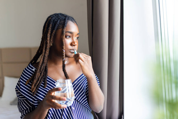 Young African woman brushing teeth with toothbrush, holding glass of water and looking in window - 写真・画像