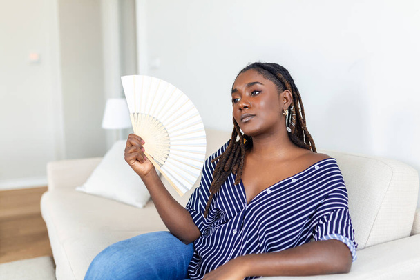 Donna afro stanca che soffre di ictus termico o caldo appartamento estivo senza aria condizionata, utilizzando ventilatore ondulato, seduto in soggiorno a casa. Surriscaldamento - Foto, immagini