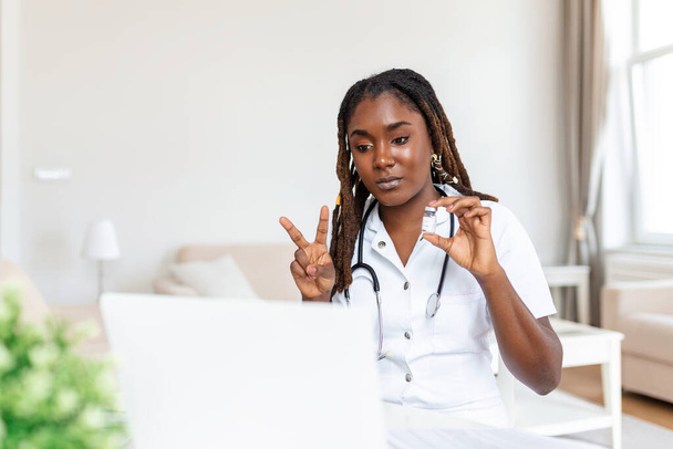 Αφρικανή ιατρός μιλάει διαδικτυακά με ασθενή, κάνει βιντεοκλήση, κοιτάζει κάμερα, νεαρή γυναίκα φοράει λευκή στολή με στηθοσκόπιο μιλώντας, συμβουλεύοντας και θεραπεύοντας έννοια - Φωτογραφία, εικόνα