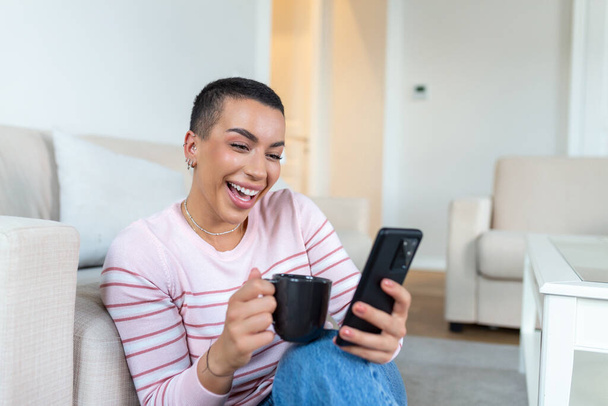 Una giovane donna in un maglione con una tazza in mano guarda il telefono mentre è seduta sul divano. - Foto, immagini