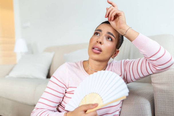 Overheated woman sit on couch at home feel warm waving with hand fan cooling down, sweating girl relax on sofa in living room hold waver suffer from heat, no air conditioner system - Photo, Image