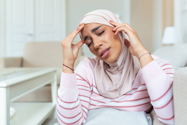 Porträt einer jungen arabischen Muslimin, die mit Kopfschmerzen und Schmerzen zu Hause auf der Couch sitzt. Frau mit Hijab leidet unter chronischen täglichen Kopfschmerzen. Traurige Frau hält ihren Kopf wegen Nebenhöhlenschmerzen - Foto, Bild