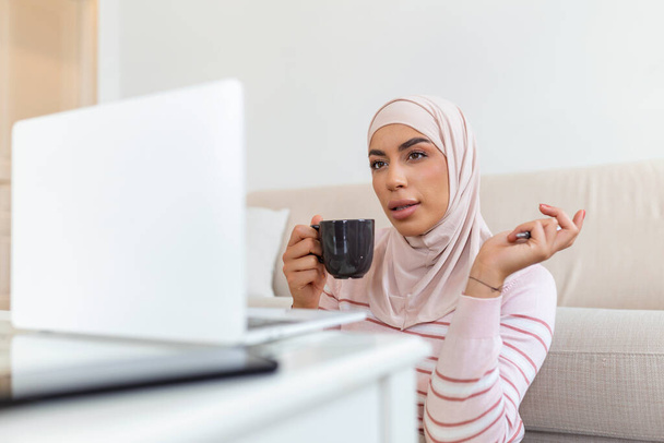 Elegancka atrakcyjna muzułmanka używająca mobilnego laptopa do wyszukiwania informacji na temat zakupów online w salonie w domu. Portret szczęśliwej kobiety pijącej kawę - Zdjęcie, obraz
