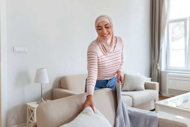 Υπέροχη νεαρή αραβίδα μουσουλμάνα γυναίκα βάζοντας μαλακά μαξιλάρια και καρό σε άνετο καναπέ, κάνοντας το σπίτι της άνετο και ζεστό - Φωτογραφία, εικόνα