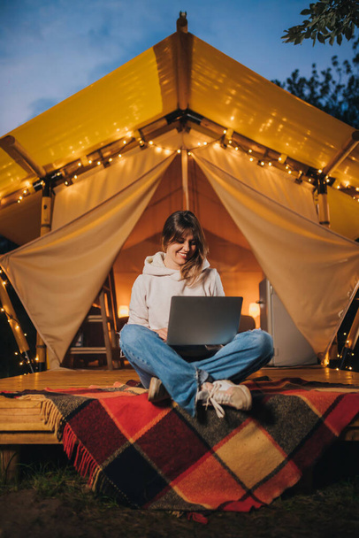 Счастливая женщина фрилансер с помощью ноутбука на уютном глампинг палатке в летний вечер. Люксовый кемпинг-шатер для отдыха на свежем воздухе. Концепция образа жизни - Фото, изображение