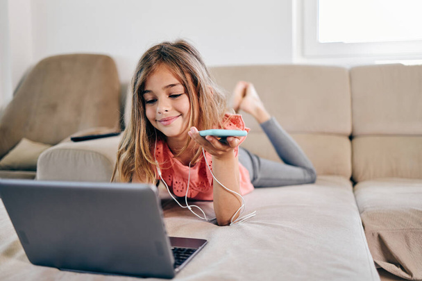 Beautiful girl lies on the living room sofa and uses a laptop and smart phone for surfing, learning and communication. She is happy and smiled. - Foto, imagen