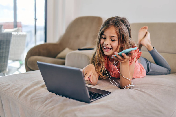 Beautiful girl lies on the living room sofa and uses a laptop and smart phone for surfing, learning and communication. She is happy and smiled. - Photo, image