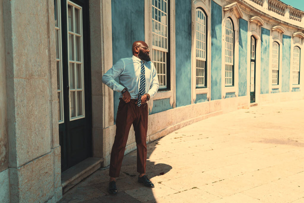 A fashionable mature bearded bald black businessman in a white shirt, glasses, and brown trousers is adjusting his belt while standing next to the door of an antique building with stained windows - Фото, зображення
