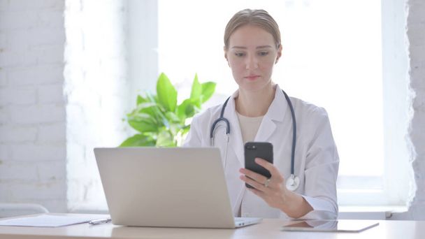 Médecin féminin utilisant Smartphone tout en utilisant un ordinateur portable dans la clinique - Photo, image
