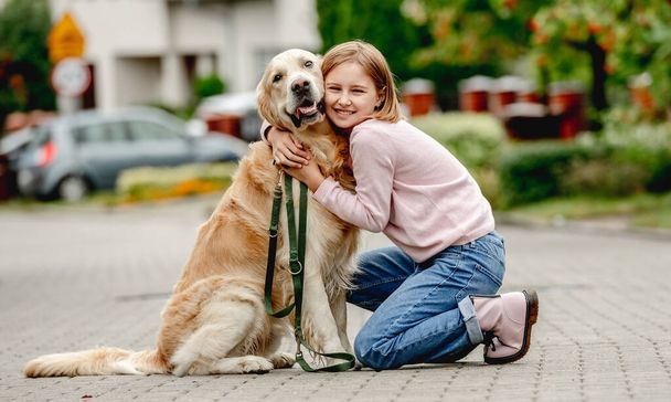 Preteen girl with golden retriever dog sitting outdoors together and looking at camera. Pretty kid child with purebred pet doggy at street - Zdjęcie, obraz