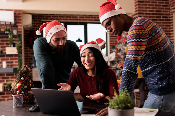 クリスマスにラップトップを使用して同僚の多民族グループは、お祝いの装飾品やクリスマスツリーで飾られたオフィスでプロジェクトに取り組んでいます。休暇中にサンタの帽子をかぶった人々がチームワークをする. - 写真・画像